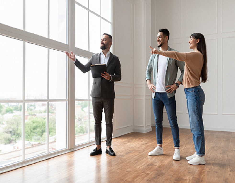 pmm Professional Real Estate Agent Showing Spacious New Apartment To Young Couple, Looking And Pointing At Beautiful View From Big Panoramic Picture Windows. Happy Man And Woman Ready to Become Homeowners